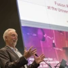 A man in a brown suit stands in front of a podium with his hands in front of him. Behind him is a screen with a presentation that reads "Fusion Research a the University of Arizona"