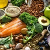Colorful spread of healthy whole foods including a raw salmon fillet, avocado, blueberries, apple, banana, kale, broccoli, cherry tomatoes, red onion, lemon, chia seeds, quinoa, beans, assorted nuts and seeds, olive oil, and various leafy greens arranged on a rustic wooden surface. A glass jar spills over with pinto beans in the background, adding texture and warmth to the nutrient-rich display.